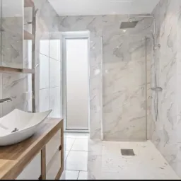 Nowoczesna łazienka z marmurowymi płytkami, kabiną prysznicową typu walk-in i asymetryczną umywalką nablatową na drewnianej szafce.