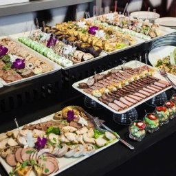 As Gastro - Bogato zastawiony stół cateringowy z różnorodnymi przekąskami: roladki mięsne, pasztety, koreczki, sałatki w szklankach, dekorowane kwiatami i ziołami, ustawione na białych tacach i paterach...