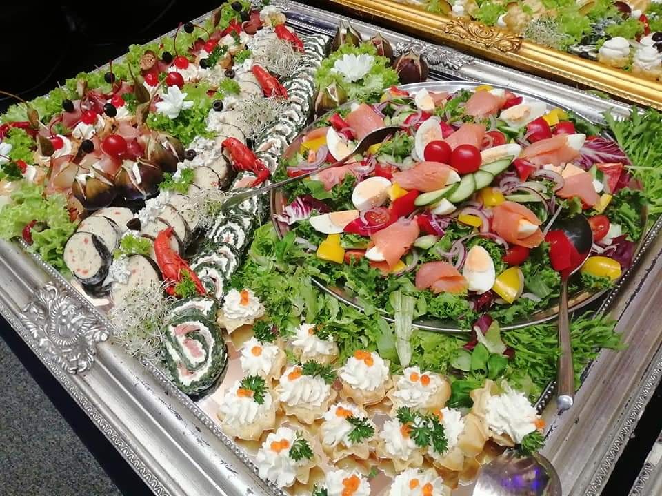 Srebrna patera z różnorodnymi przekąskami: sałatka z łososiem, roladki szpinakowe, tartaletki z kremem i ikrą, koreczki z figami i szynką, dekorowane zieleniną i kwiatami.