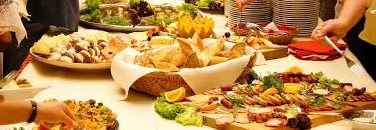 Stół bufetowy z różnorodnymi przekąskami: wędliny, sery, pieczywo w koszyku, sałatki; widoczne ręce nakładające jedzenie, sterta talerzy w tle.