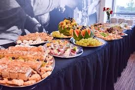 Stół cateringowy z różnorodnymi przekąskami: kanapeczki, sałatki, owoce (arbuzy rzeźbione w kształcie koszyczków), czarny obrus, w tle grafika na ścianie.