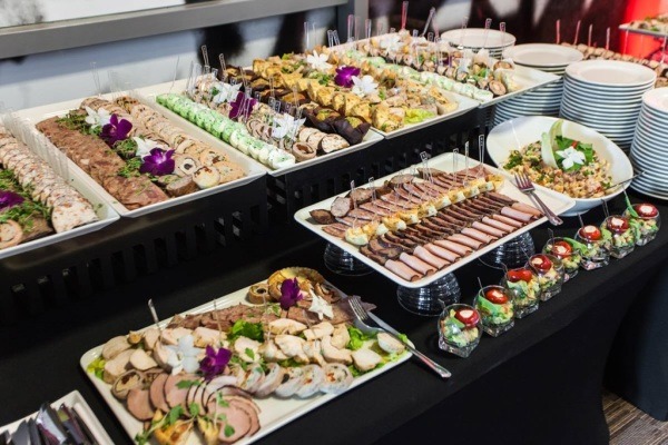 Bogato zastawiony stół cateringowy z różnorodnymi przekąskami: roladki mięsne, pasztety, koreczki, sałatki w szklankach, dekorowane kwiatami i ziołami, ustawione na białych tacach i paterach...