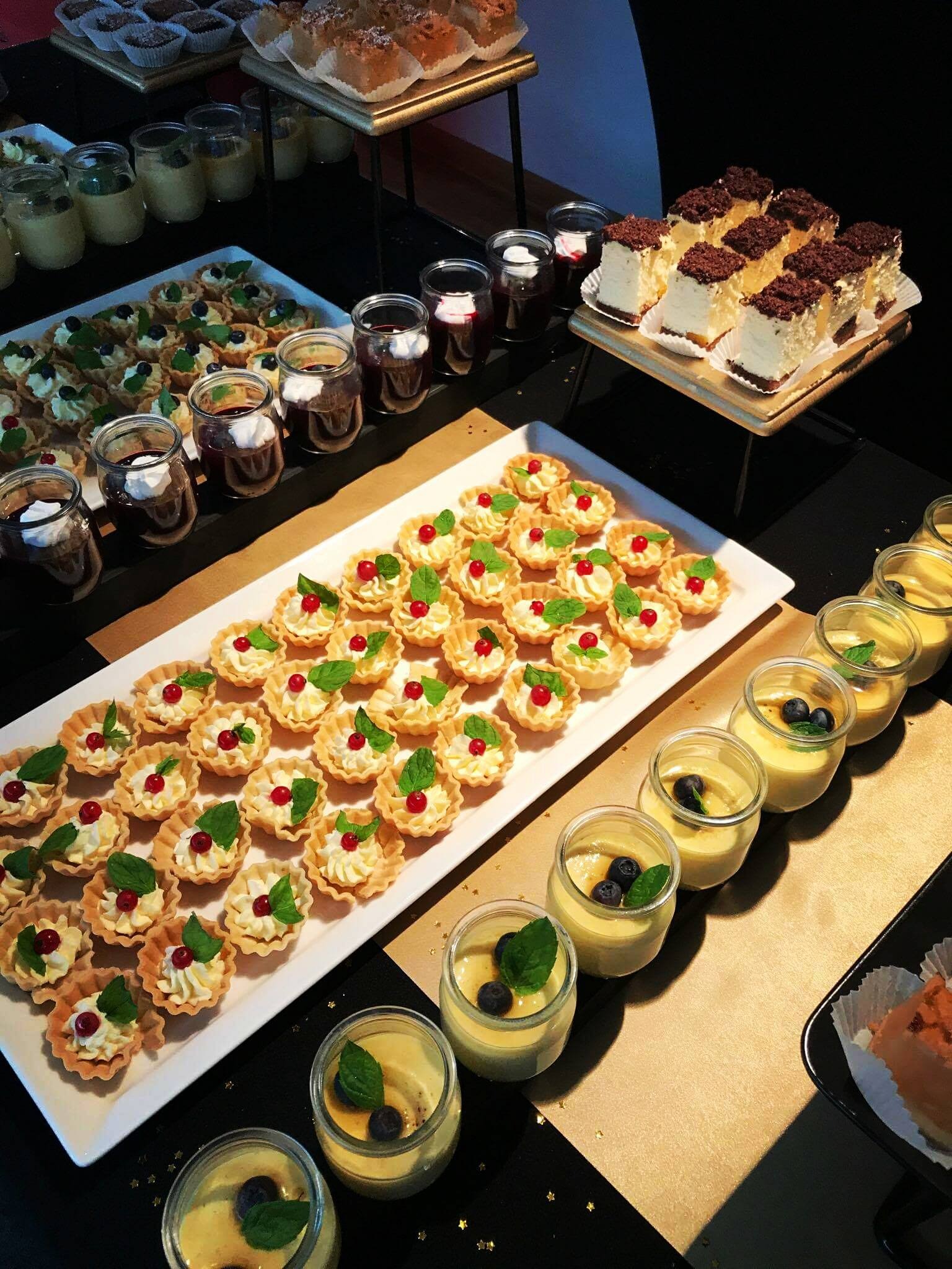 Elegancki bufet z różnorodnymi deserami: tartaletki z kremem i czerwoną porzeczką, puddingi w słoiczkach z borówkami i miętą, ciasta z czekoladową posypką, całość na czarnym obrusie ze złotymi...