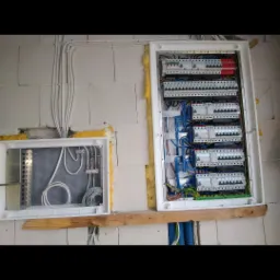 Dwie skrzynki elektryczne zamontowane na ścianie z bloczków, skrzynka po prawej stronie zawiera rozdzielnicę z bezpiecznikami i okablowaniem, skrzynka po lewej stronie zawiera instalację...