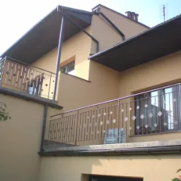 Dwupiętrowy dom z beżową elewacją, balkonami z metalowymi balustradami o geometrycznym wzorze rombów, widoczne okna PCV i rynny.