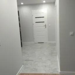 Korytarz po remoncie z szarą podłogą imitującą marmur i panelami imitującymi drewno, białe drzwi z trzema czarnymi paskami i oświetlenie punktowe w suficie.