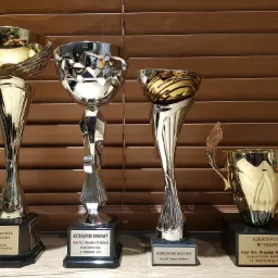 Cztery błyszczące puchary z napisami 'Kierownik Budowy' i nazwiskiem 'Bogdan Stodziesz', reprezentujące sukcesy firmy z Ostrowa Wielkopolskiego w branży budowlanej, ustawione na tle drewnianych...