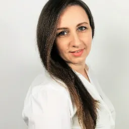 Portret uśmiechniętej kobiety z długimi brązowymi włosami w białej koszuli na jasnym tle.