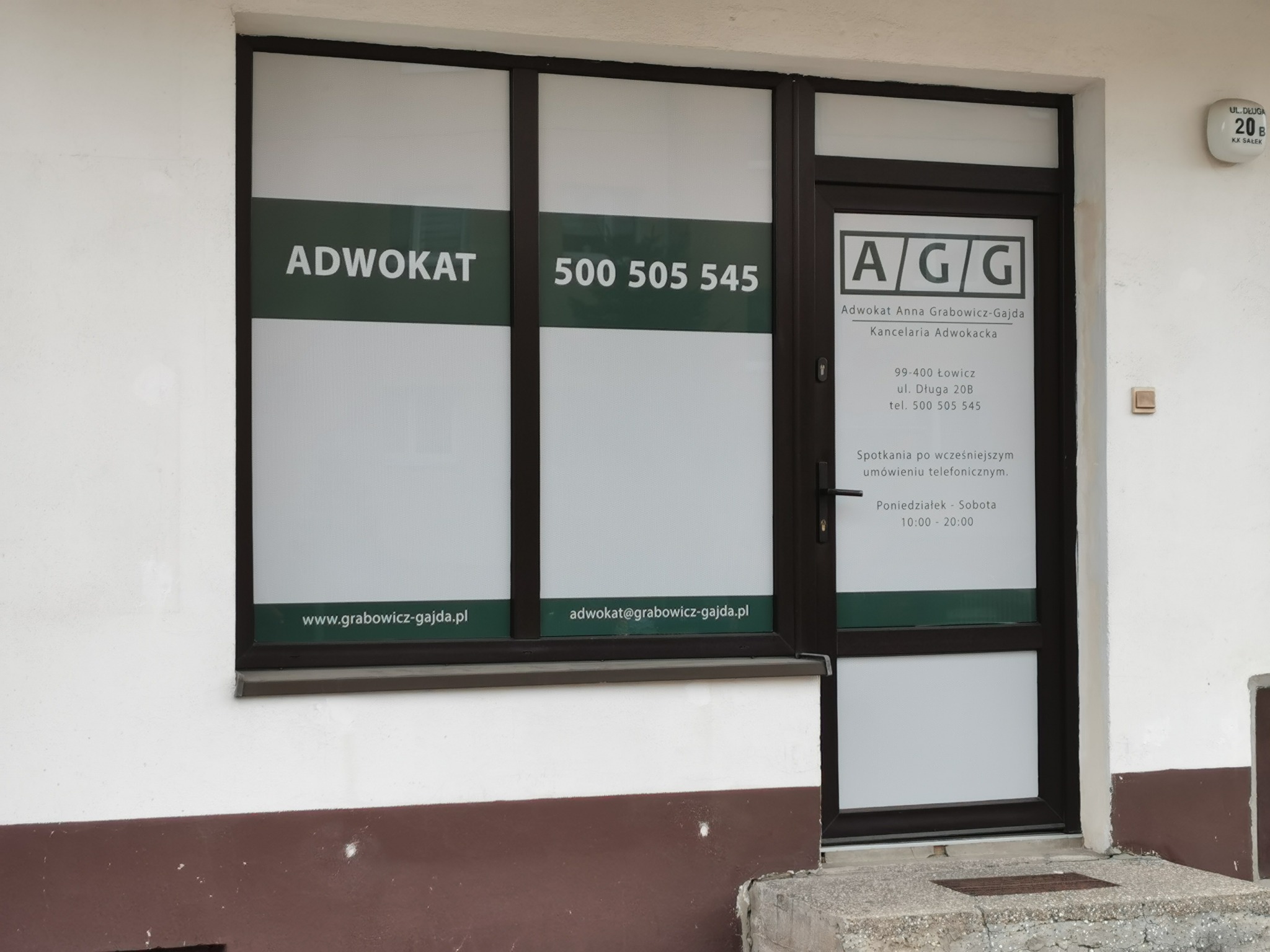 Fasada budynku z szyldem kancelarii adwokackiej, widoczne okna z napisem 'Adwokat' i numerem telefonu, drzwi wejściowe z informacjami o kancelarii i godzinach otwarcia.