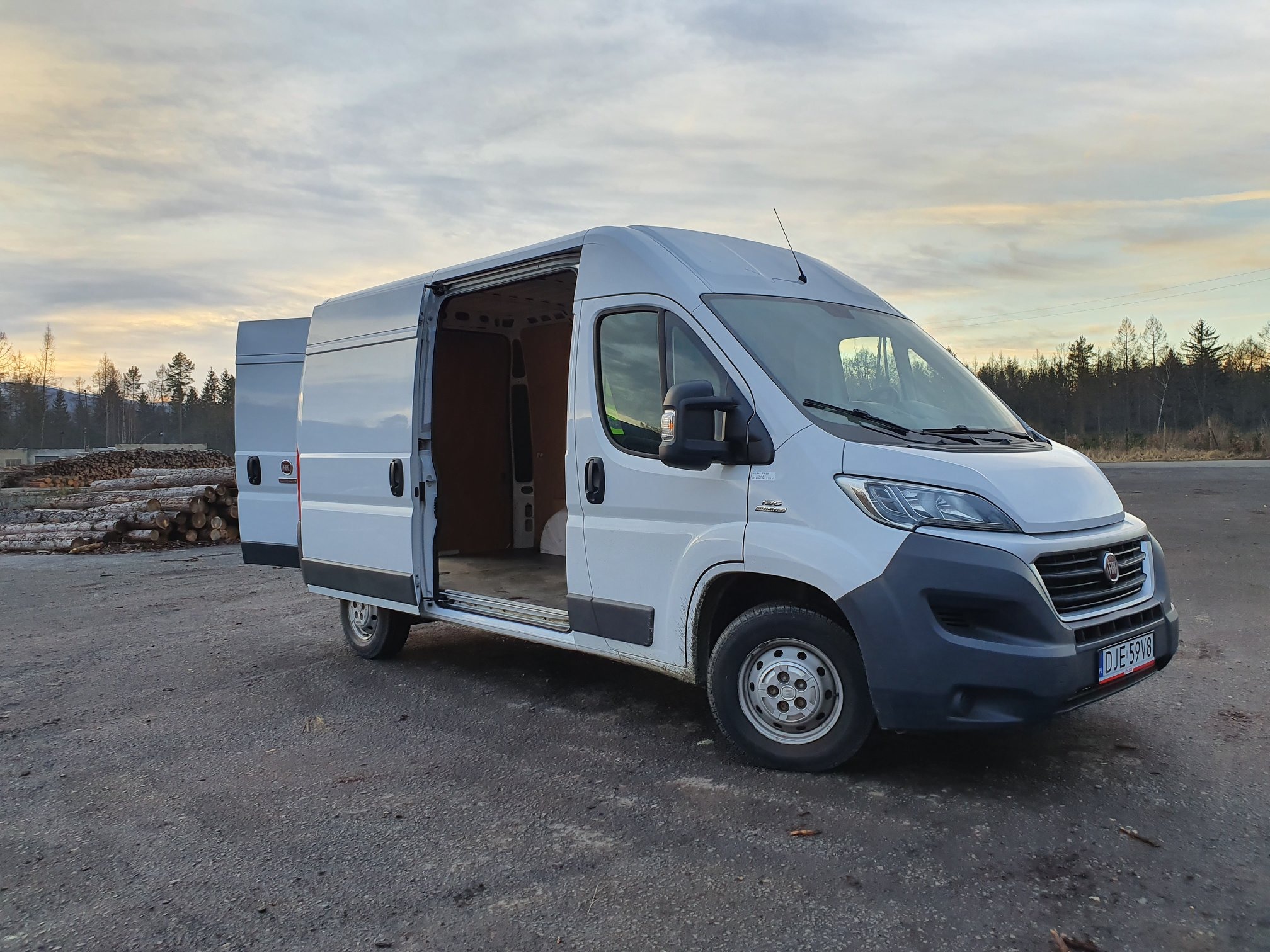 Biały bus dostawczy marki Fiat z otwartymi drzwiami bocznymi i tylnymi, zaparkowany na szarym asfalcie z widocznymi stosem pni drzew w tle.