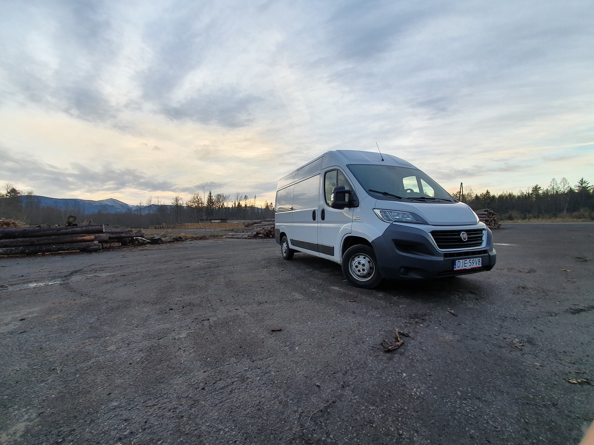 Biały bus dostawczy Fiat Ducato zaparkowany na szarym, asfaltowym placu z widokiem na stertę drewna i zalesione wzgórza w tle, pochmurne niebo.