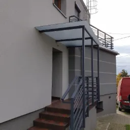 Zewnętrzne schody z terakotowymi płytkami prowadzące do drzwi wejściowych, zabezpieczone metalową balustradą i zadaszeniem z przezroczystego szkła.