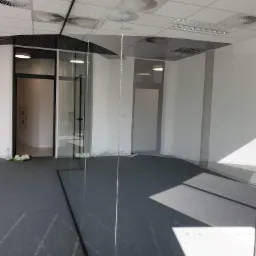 Szklana ściana działowa w nowoczesnym biurze z szarą wykładziną, widoczne białe ślady po montażu i butelki po płynie czyszczącym, czarne ramy drzwi i okien, białe ściany, sufit z wentylacją.