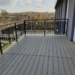 Nowoczesny balkon z szarą kompozytową podłogą i szklaną balustradą z czarnymi elementami, widok na zabudowania miejskie w tle.