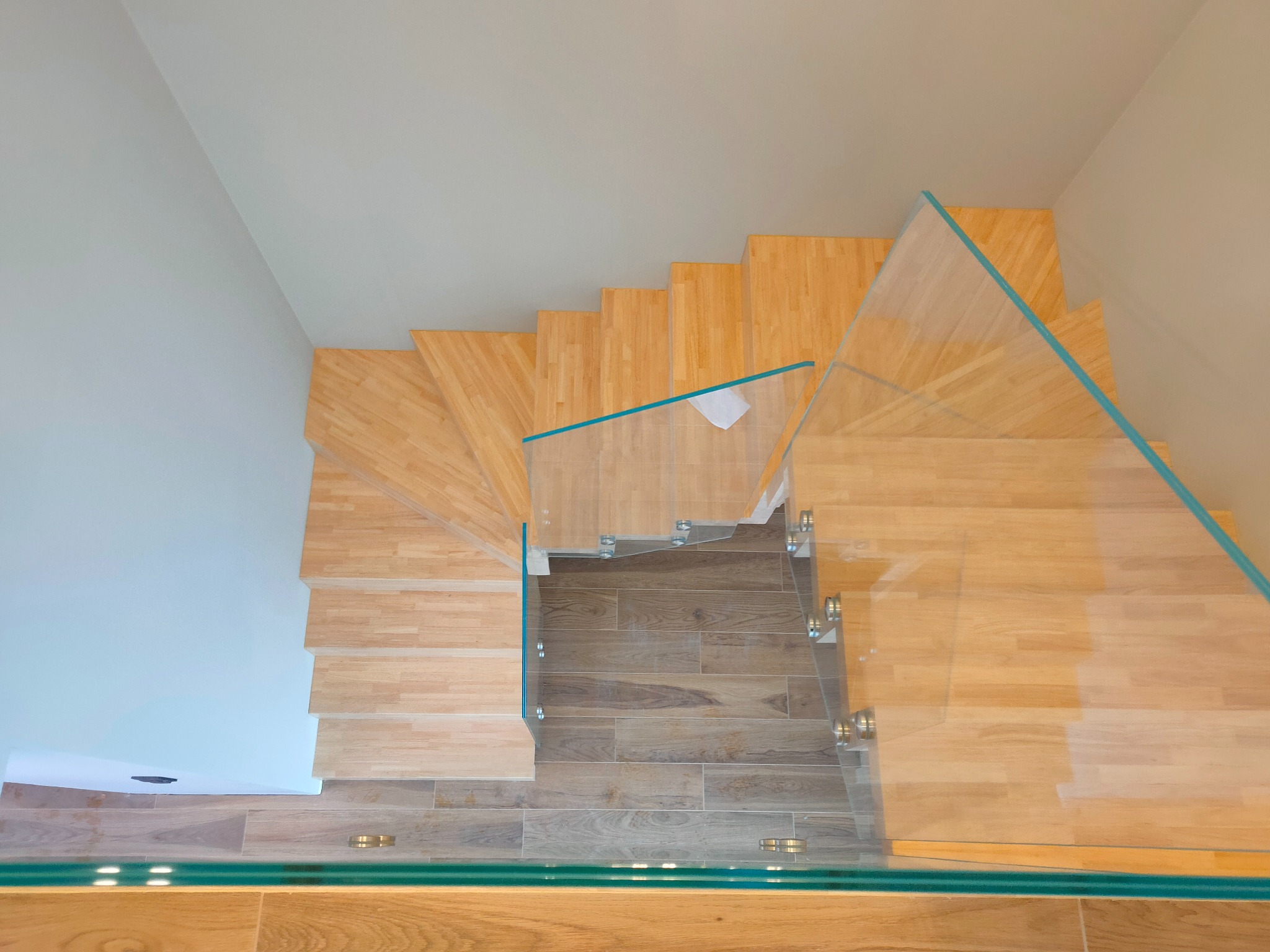 Widok z góry na jasne, drewniane stopnie z przezroczystą szklaną balustradą zamontowaną na metalowych uchwytach, prowadzące na brązową podłogę w Bytomiu.