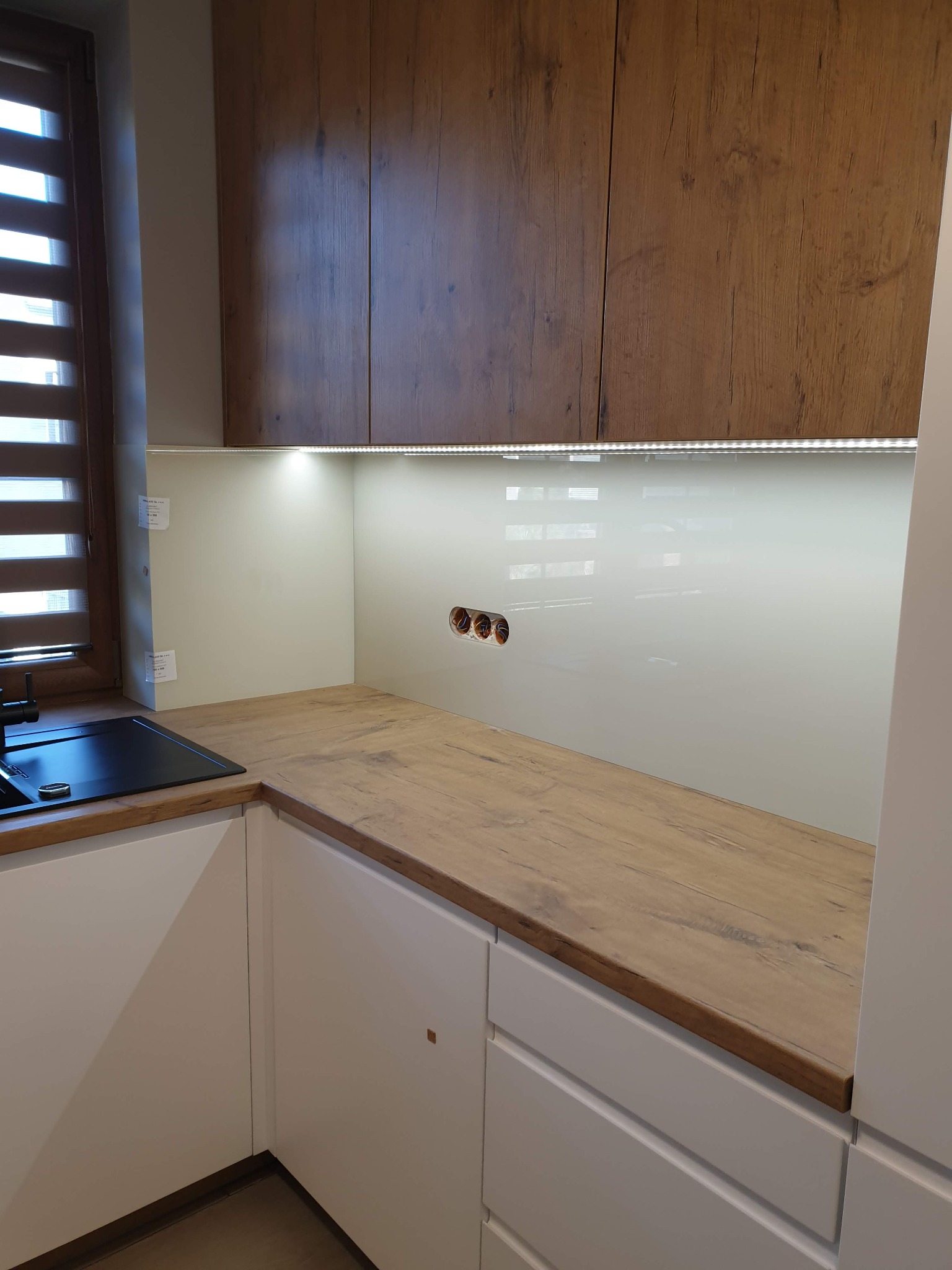 Nowoczesna kuchnia z drewnianymi szafkami górnymi, białymi szafkami dolnymi i blatem o fakturze drewna, podświetlona listwą LED, z widocznym oknem z żaluzjami poziomymi i czarnym zlewem.