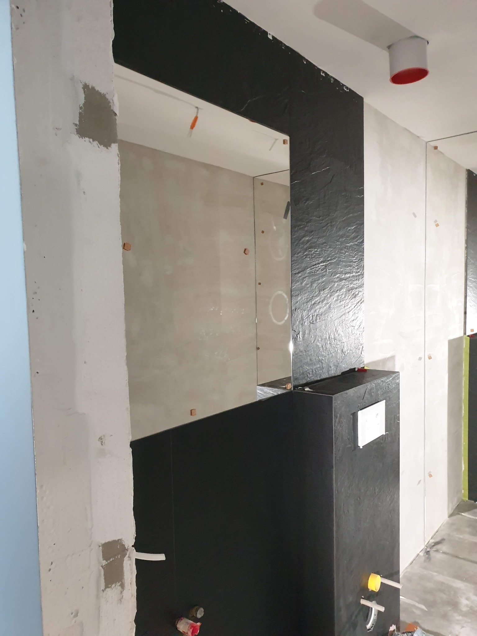 Wnętrze łazienki w trakcie prac wykończeniowych, widoczne czarne i białe płytki o nieregularnej fakturze, fragmenty ścian w stanie surowym, elementy instalacji hydraulicznej i podłączeń, oraz rury...