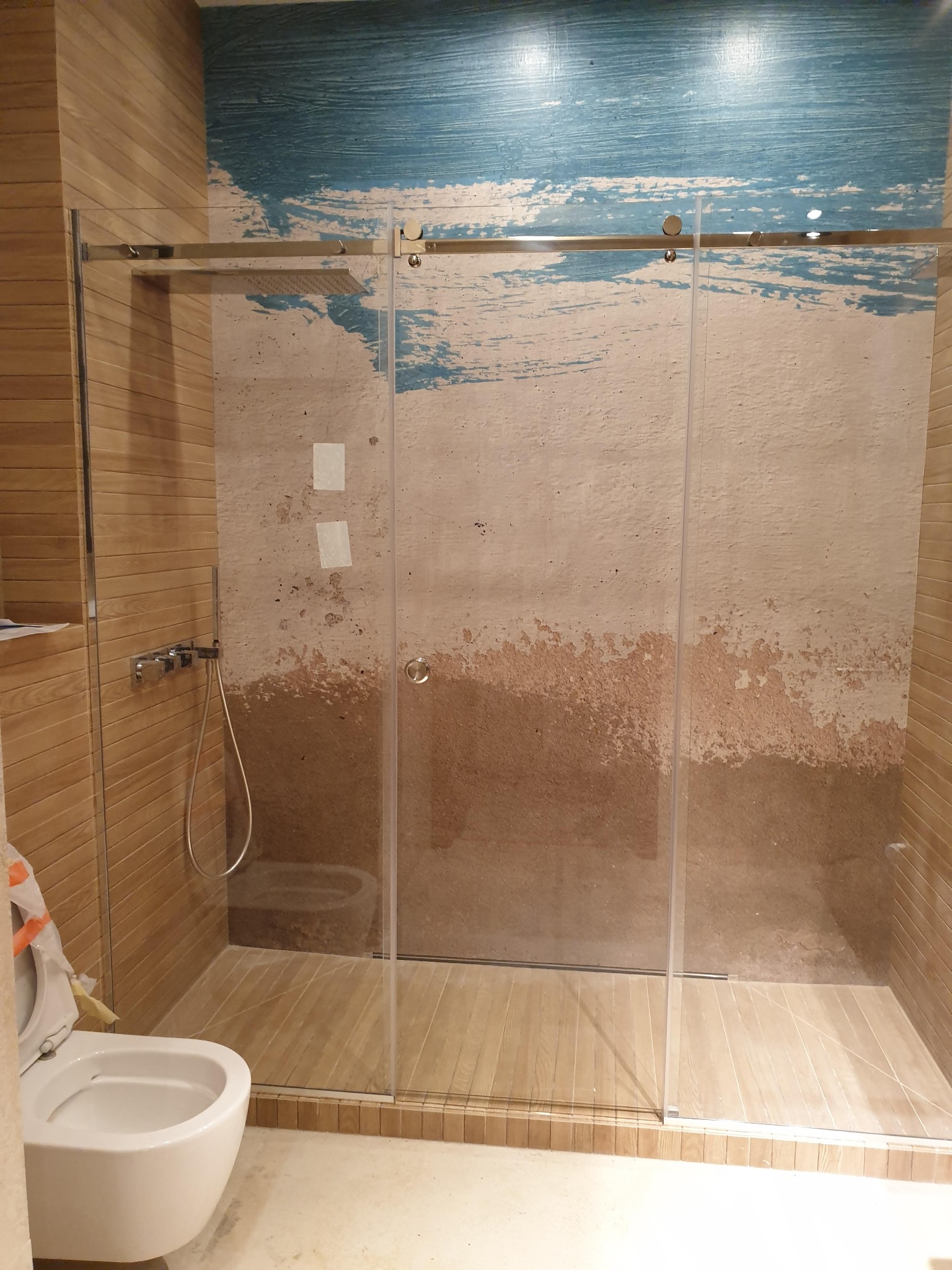 Szklana kabina prysznicowa z przesuwnymi drzwiami, zamontowana w łazience z drewnopodobnymi płytkami i dekoracyjną ścianą imitującą piasek i wodę, widoczny fragment toalety.