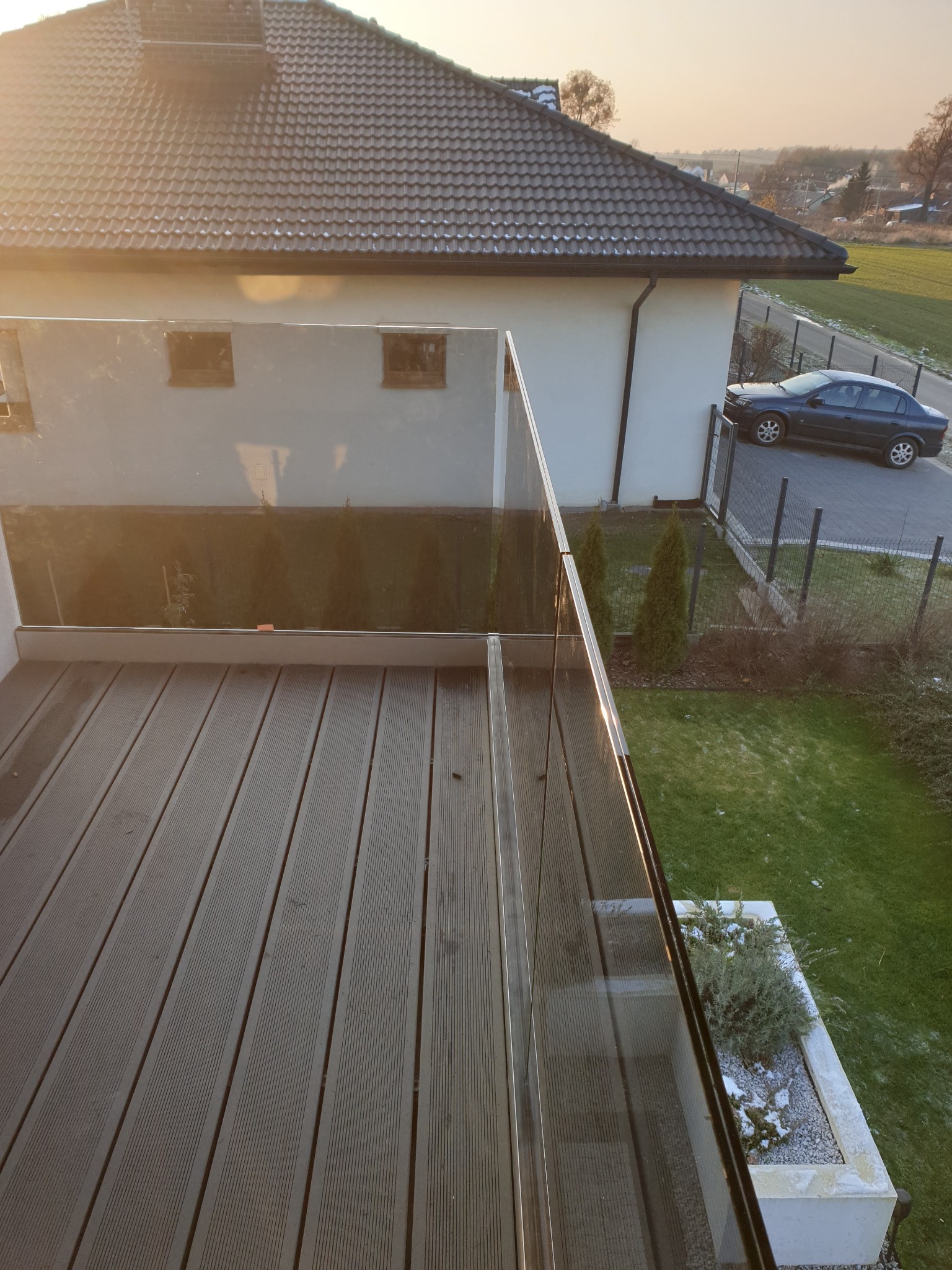 Zewnętrzny taras z desek kompozytowych, zabezpieczony szklaną balustradą, widok z góry na ogród z roślinami i dom z ciemnym dachem w tle, wczesne popołudnie.