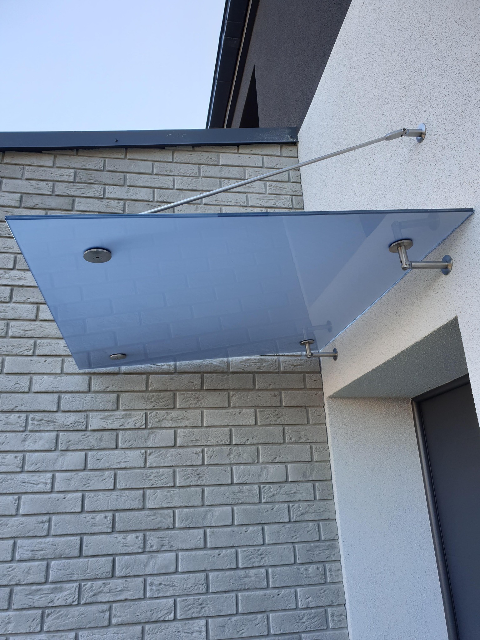 Szklany daszek nad wejściem do domu, wsparty na metalowych wspornikach, z widoczną fakturą ceglanej ściany z lewej strony.