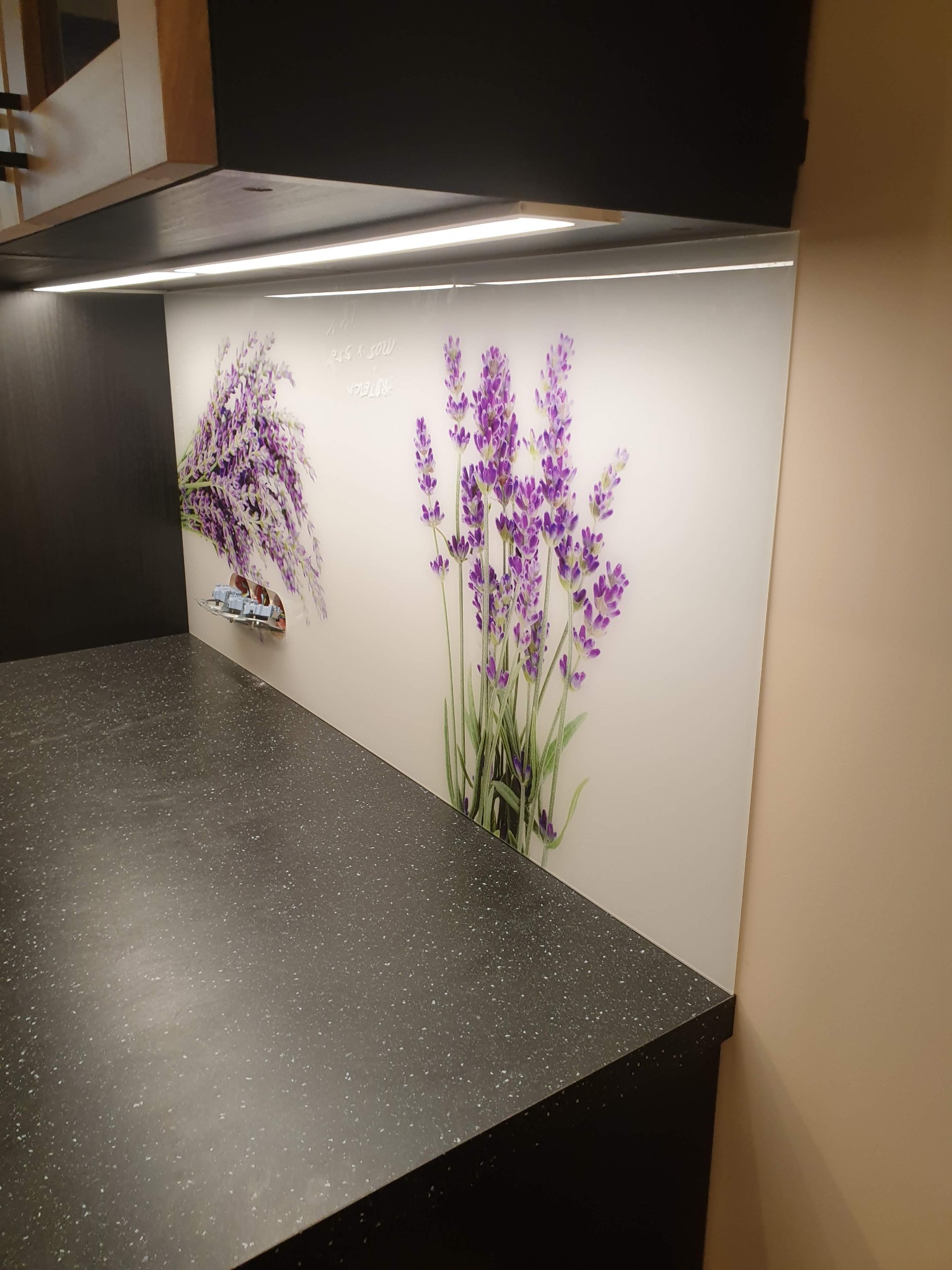 Kuchenny blat z ciemnego kamienia, nad którym znajduje się panel ścienny z motywem lawendy i widocznymi gniazdkami elektrycznymi.