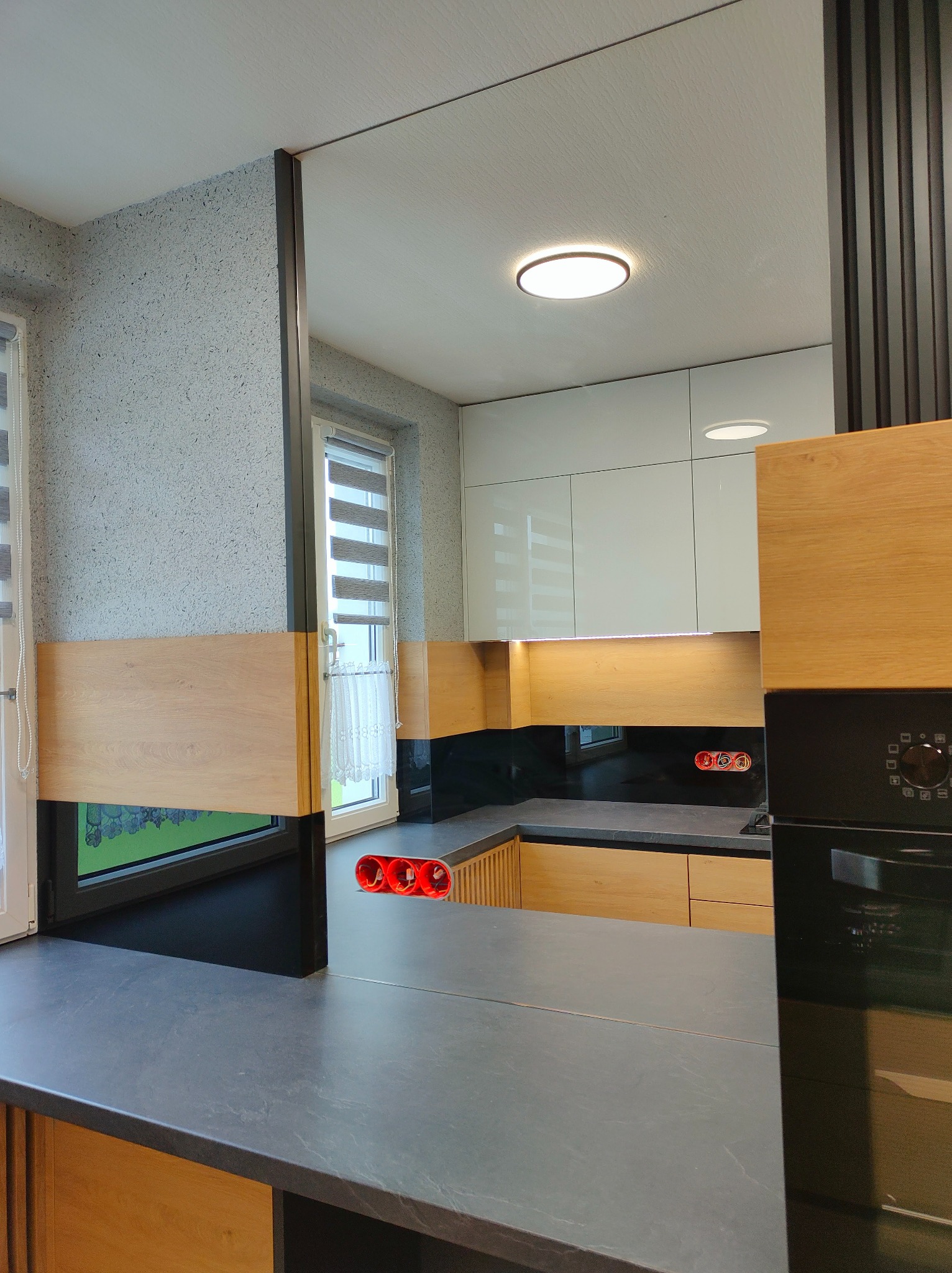 Nowoczesna kuchnia z drewnianymi i białymi szafkami, czarnym blatem i oknem z roletami w tle, widok z perspektywy blatu, w tle widoczne czerwone gniazdka.