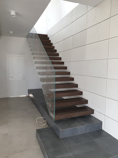 Nowoczesne schody z drewnianymi stopniami, szklaną balustradą i białymi płytami ściennymi, oświetlone światłem dziennym z góry.