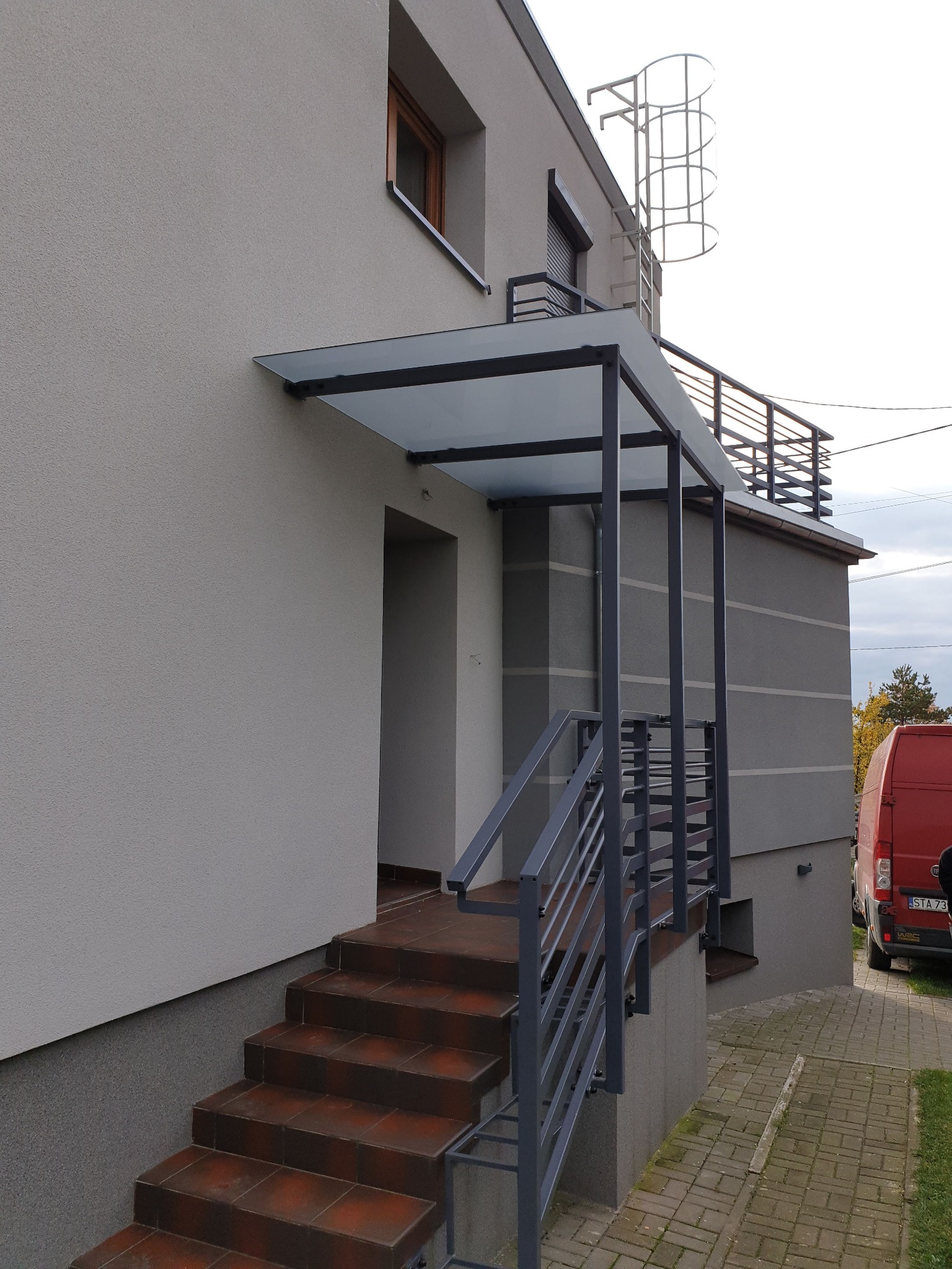 Zewnętrzne schody z terakotowymi płytkami prowadzące do drzwi wejściowych, zabezpieczone metalową balustradą i zadaszeniem z przezroczystego szkła.