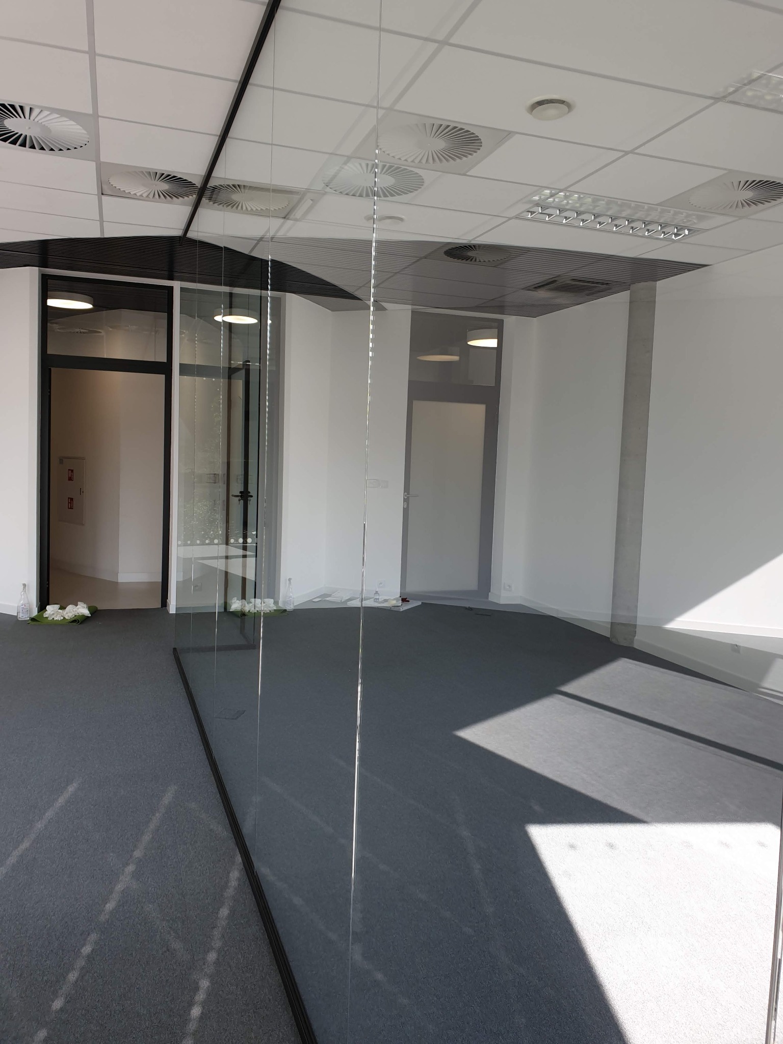 Szklana ściana działowa w nowoczesnym biurze z szarą wykładziną, widoczne białe ślady po montażu i butelki po płynie czyszczącym, czarne ramy drzwi i okien, białe ściany, sufit z wentylacją.