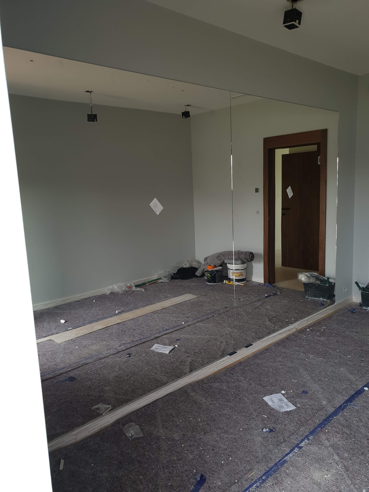 Wnętrze pokoju w trakcie prac wykończeniowych z dużym lustrem na ścianie, zabezpieczoną podłogą, widocznymi narzędziami i otwartymi drzwiami w tle.