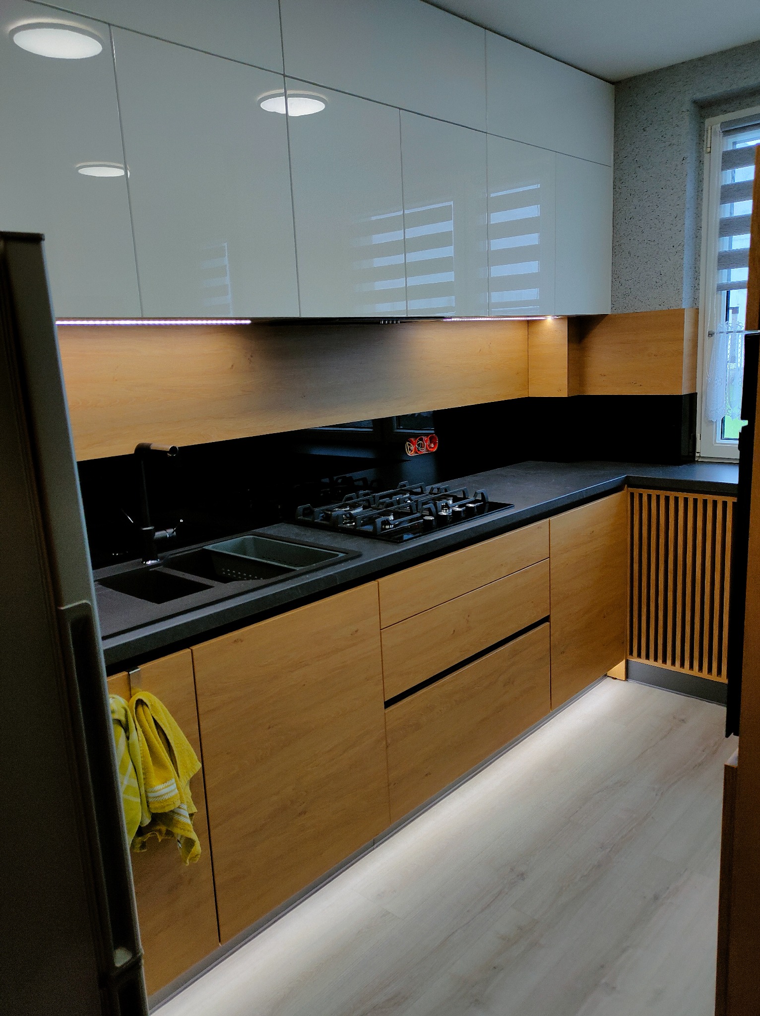 Nowoczesna kuchnia z drewnianymi szafkami dolnymi, czarnym blatem i białymi szafkami górnymi, oświetlona listwami LED pod szafkami wiszącymi i przy podłodze, z widocznym fragmentem lodówki i żółtą...