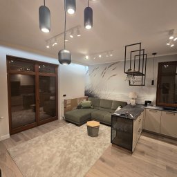 GadMar-Bud - Jasne wnętrze po wykończeniu: drewniane okna, sofa z poduszką, dywan z wzorem, kuchnia z czarnym blatem i designerskie oświetlenie sufitowe.