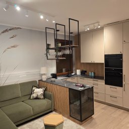 GadMar-Bud - Nowoczesna kuchnia z wyspą i barkiem na wino,  w odcieniach beżu i czerni, z zieloną sofą w tle. Na ścianie fototapeta z motywem trawy.