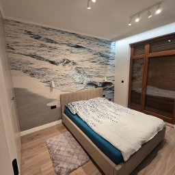 GadMar-Bud - Sypialnia po remoncie: łóżko z beżową ramą, pościel w błękitne wzory, fototapeta z motywem morza i drewniana podłoga. Nowoczesne oświetlenie punktowe.
