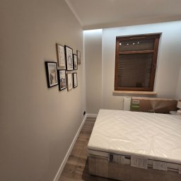 GadMar-Bud - Wykończony pokój z łóżkiem i galerią obrazków w Pruszczu Gdańskim. Ściany w odcieniach beżu, drewniana podłoga i okno z żaluzjami. Nowoczesne oświetlenie.