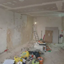 Pok&oacute;j w trakcie prac przygotowawczych do położenia gładzi: ściany z widocznymi ubytkami i fragmentami starej farby, sufit z płyt kartonowo-gipsowych, rozstawiona drabina, narzędzia i materiały...
