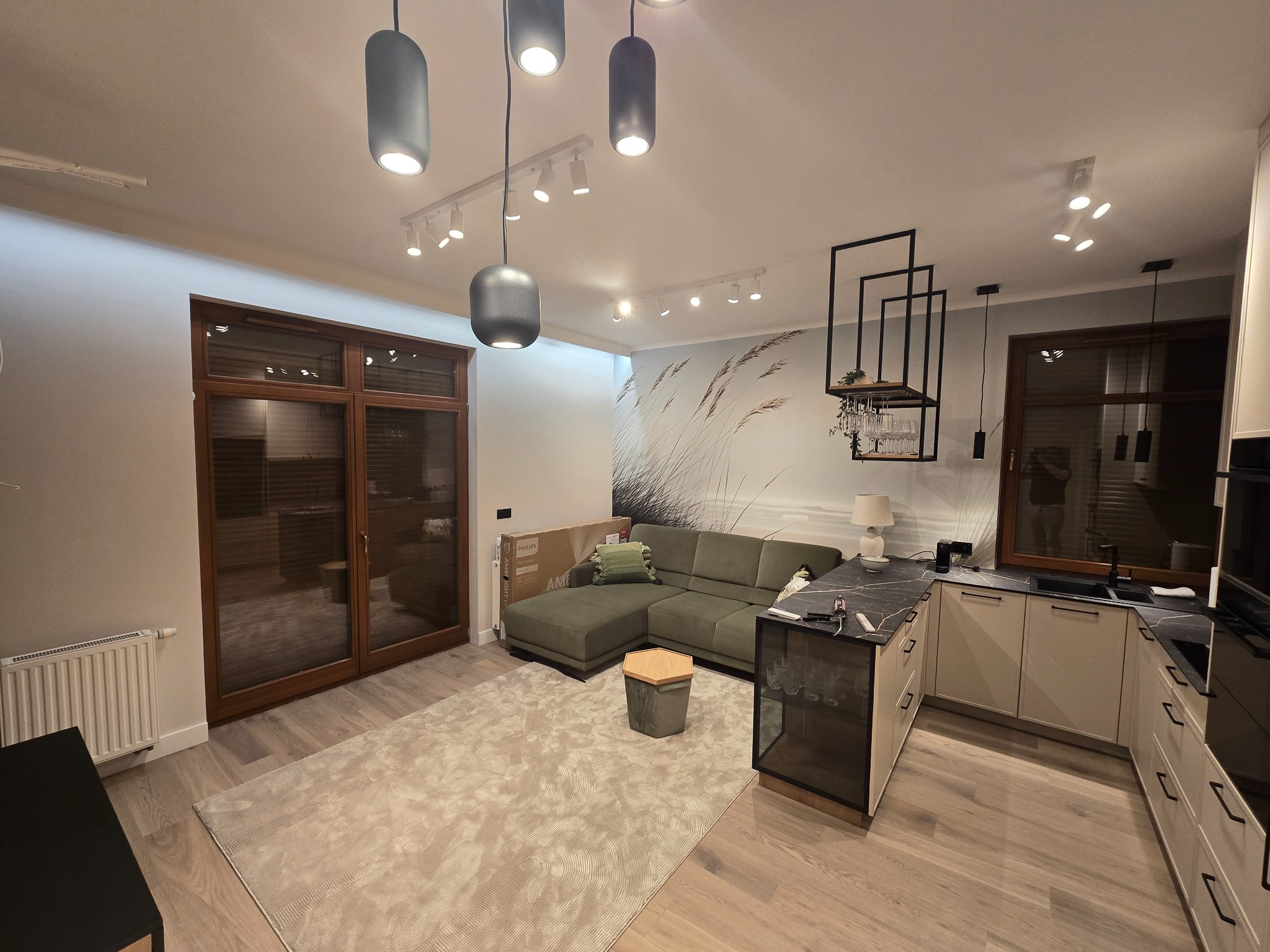 Jasne wnętrze po wykończeniu: drewniane okna, sofa z poduszką, dywan z wzorem, kuchnia z czarnym blatem i designerskie oświetlenie sufitowe.