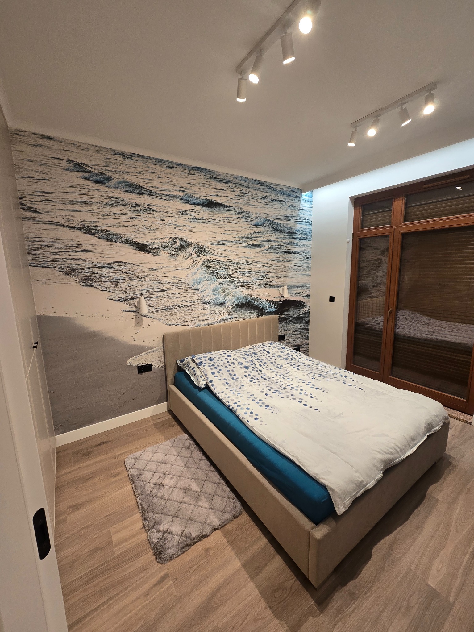 Sypialnia po remoncie: łóżko z beżową ramą, pościel w błękitne wzory, fototapeta z motywem morza i drewniana podłoga. Nowoczesne oświetlenie punktowe.