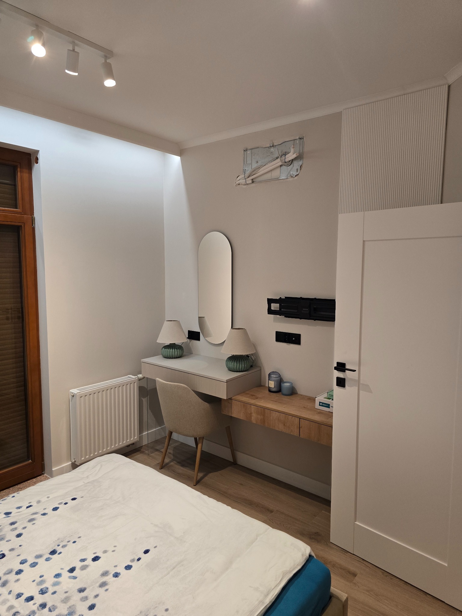 Nowoczesna sypialnia z toaletką, owalnym lustrem i minimalistycznymi lampkami. Na ścianie widoczne elementy instalacji klimatyzacji i uchwyt do TV.