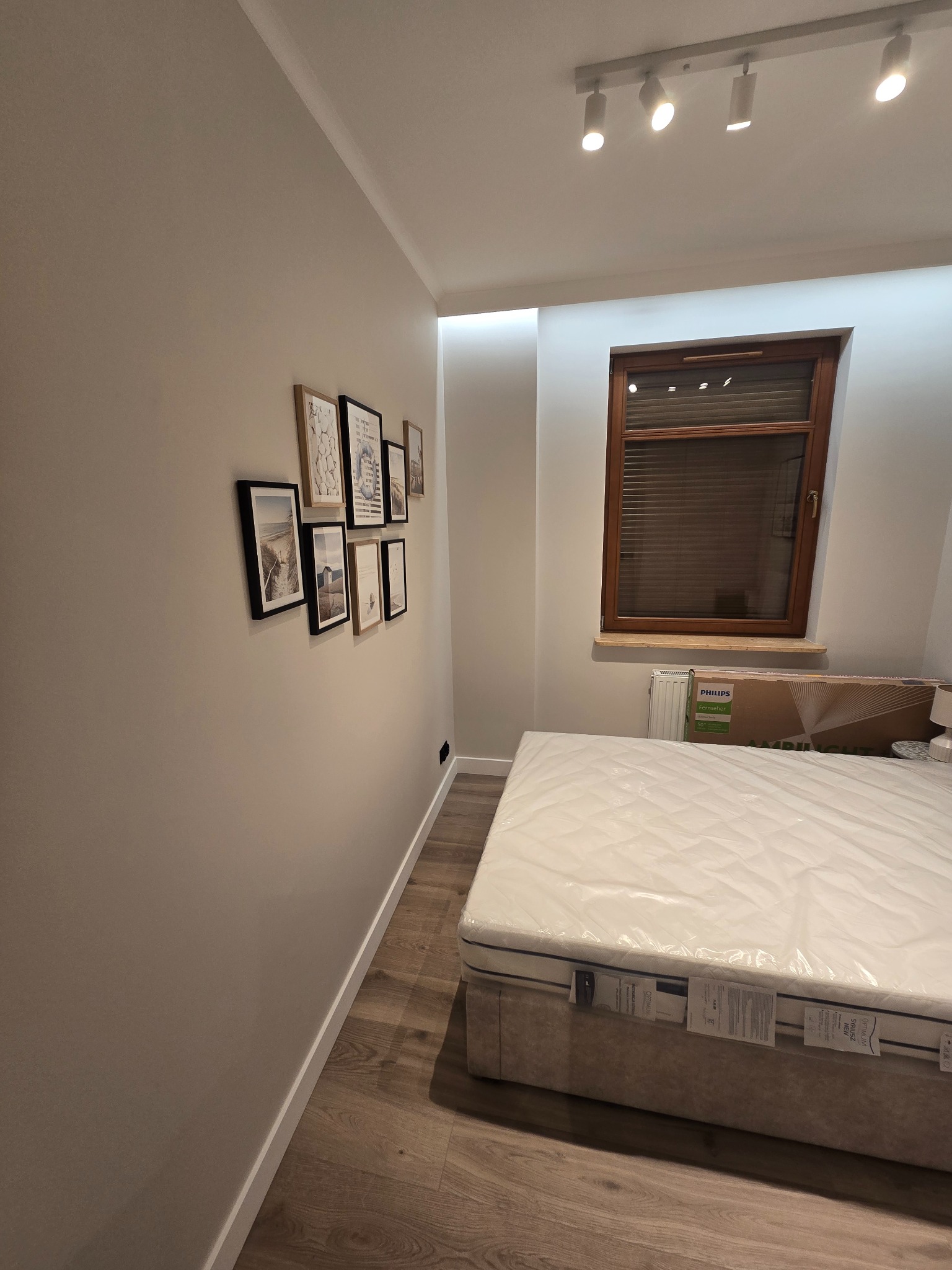 Wykończony pokój z łóżkiem i galerią obrazków w Pruszczu Gdańskim. Ściany w odcieniach beżu, drewniana podłoga i okno z żaluzjami. Nowoczesne oświetlenie.