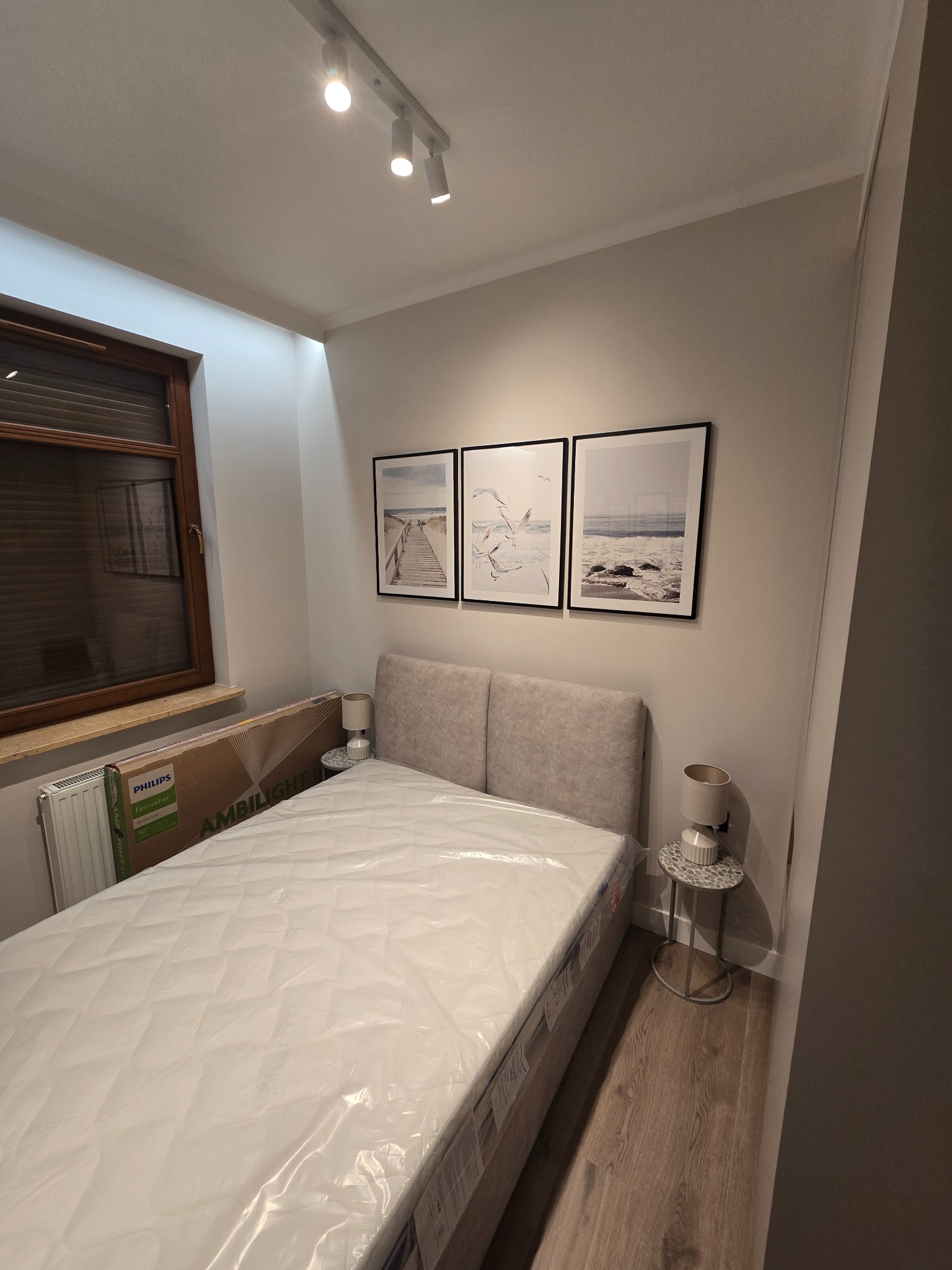 Jasna sypialnia z łóżkiem w folii, drewnianą podłogą i trzema obrazami w czarnych ramkach nad wezgłowiem. Obok łóżka stoliki z lampkami. Przy oknie karton po telewizorze.