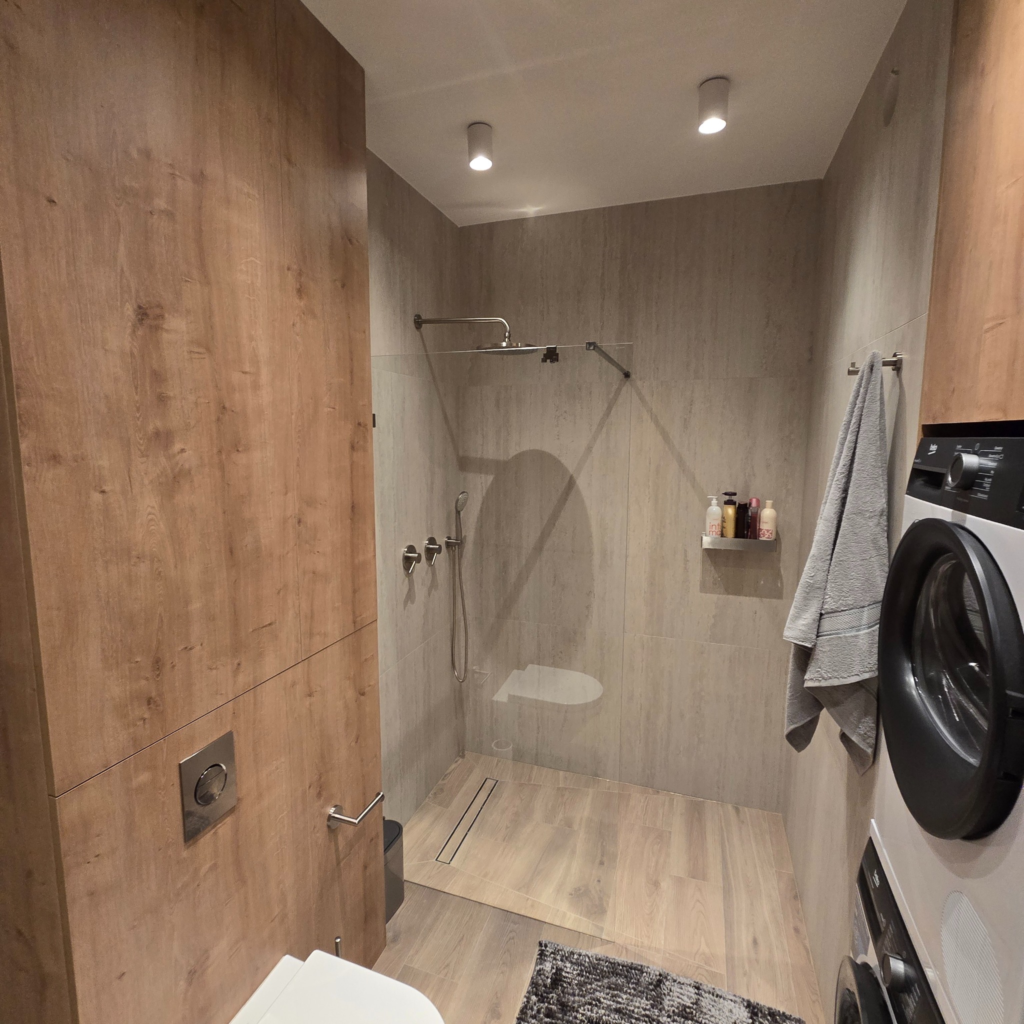 Nowoczesna łazienka po remoncie: kabina prysznicowa ze szklaną ścianą, drewnopodobne panele ścienne, pralka i minimalistyczny wystrój. Widok z perspektywy.
