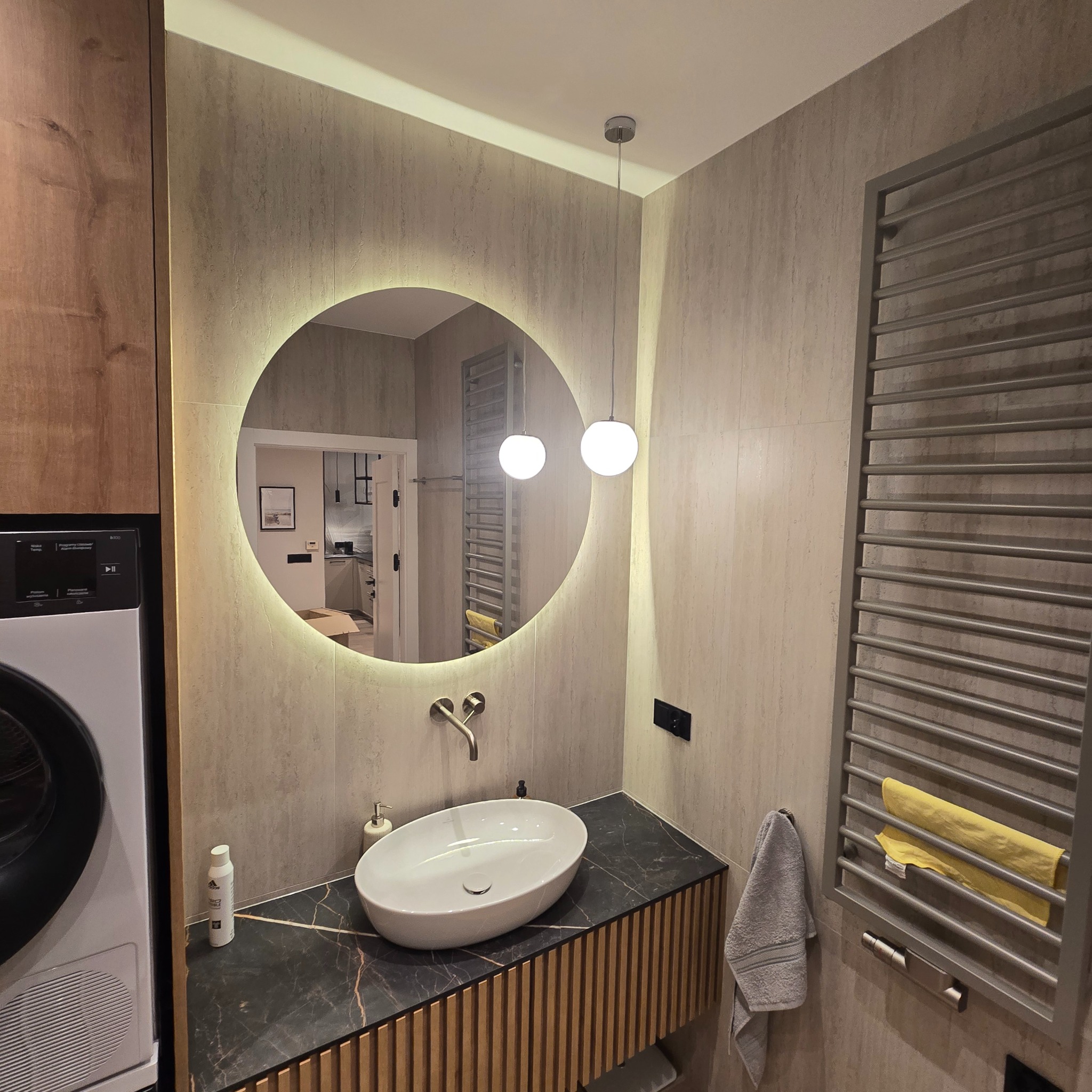 Nowoczesna łazienka z okrągłym lustrem podświetlanym LED, umywalką nablatową na szafce z drewnianymi lamelami i grzejnikiem drabinkowym z ręcznikami.