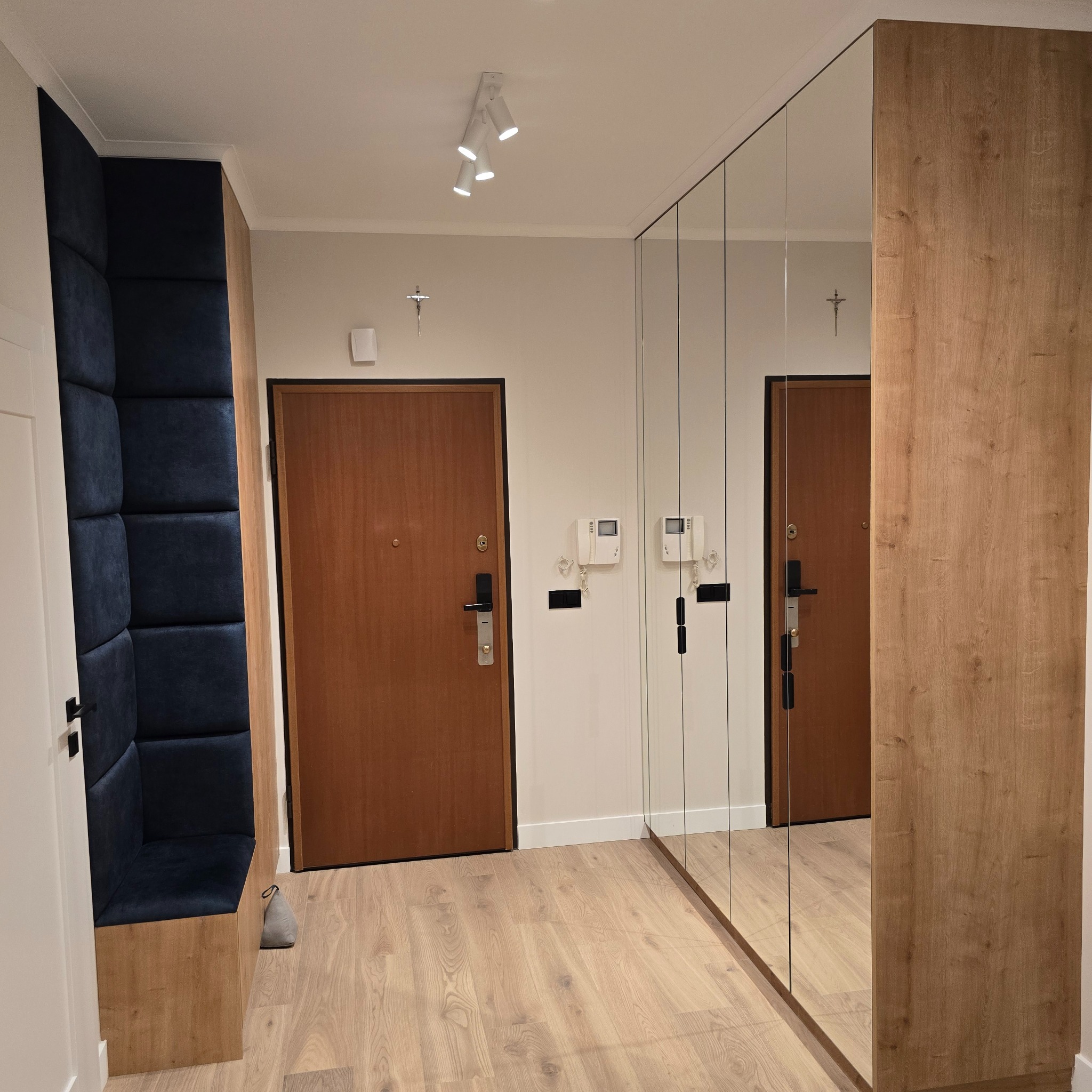 Korytarz po metamorfozie: drewniana podłoga, lustrzana szafa wnękowa i tapicerowane siedzisko w kolorze granatu. Nowoczesne oświetlenie punktowe.
