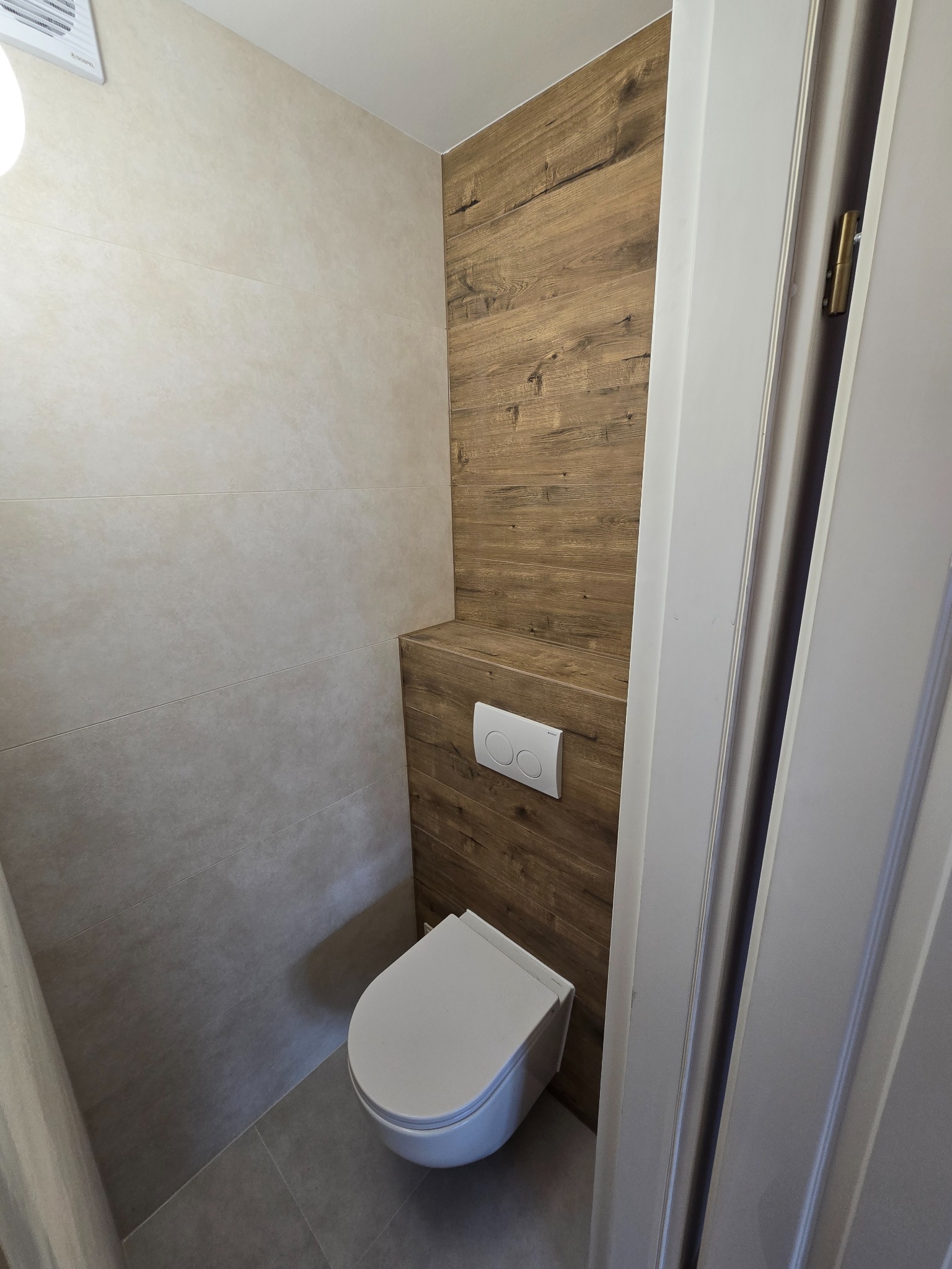 Nowoczesna toaleta z drewnianą ścianą i beżowymi płytkami. Widok na podwieszaną miskę WC i częściowo otwarte drzwi. Minimalistyczny design.