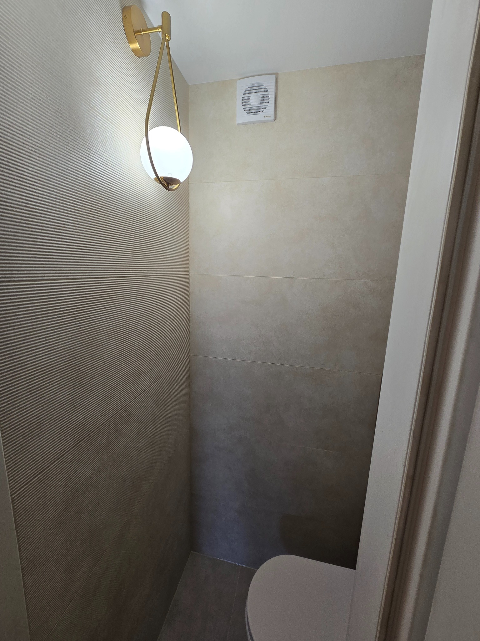 Wykończona łazienka z beżowymi płytkami, ozdobną ścianą z fakturą i nowoczesnym oświetleniem. Widoczny fragment toalety i wentylator.