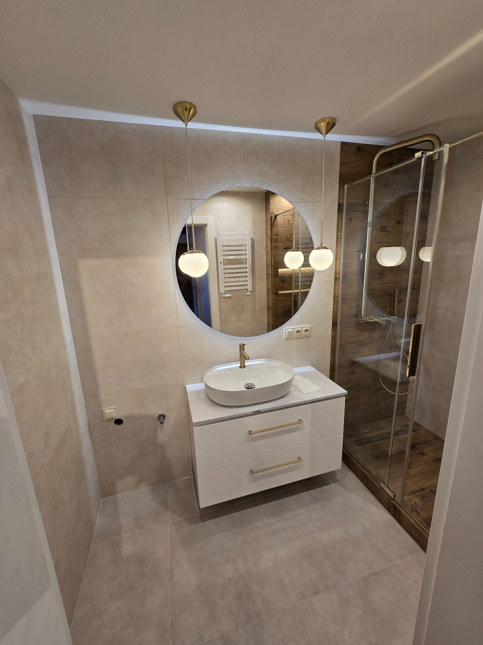 Wykończona łazienka z okrągłym lustrem LED, białą szafką z złotymi uchwytami i kabiną prysznicową z drewnianymi akcentami, utrzymana w stonowanej kolorystyce.
