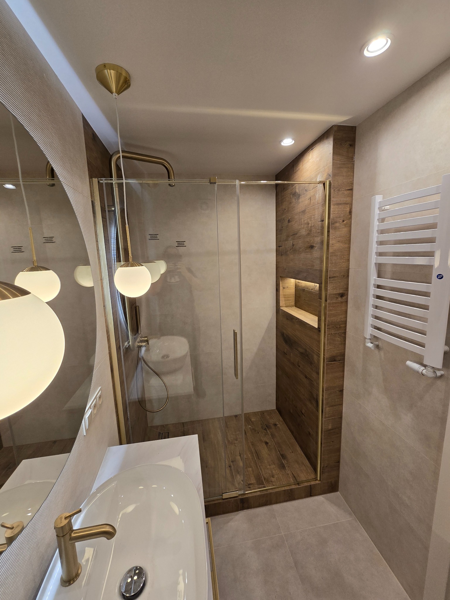 Nowoczesna łazienka z kabiną prysznicową typu walk-in, drewnianymi akcentami i złotymi detalami. Widoczna umywalka z okrągłym lustrem i grzejnik.
