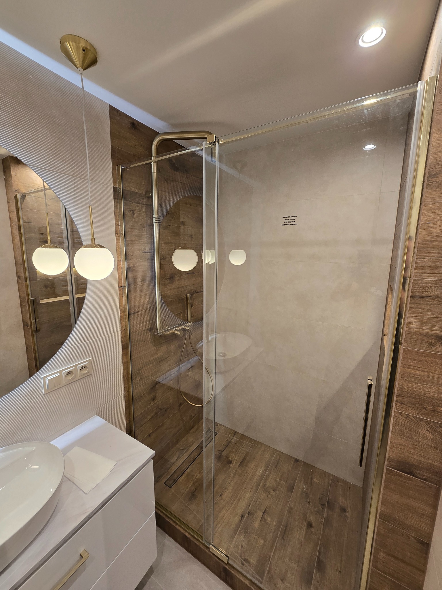 Nowoczesna kabina prysznicowa z elementami w kolorze złotym, drewnopodobne płytki na ścianach i podłodze, okrągłe lustro z lampami.