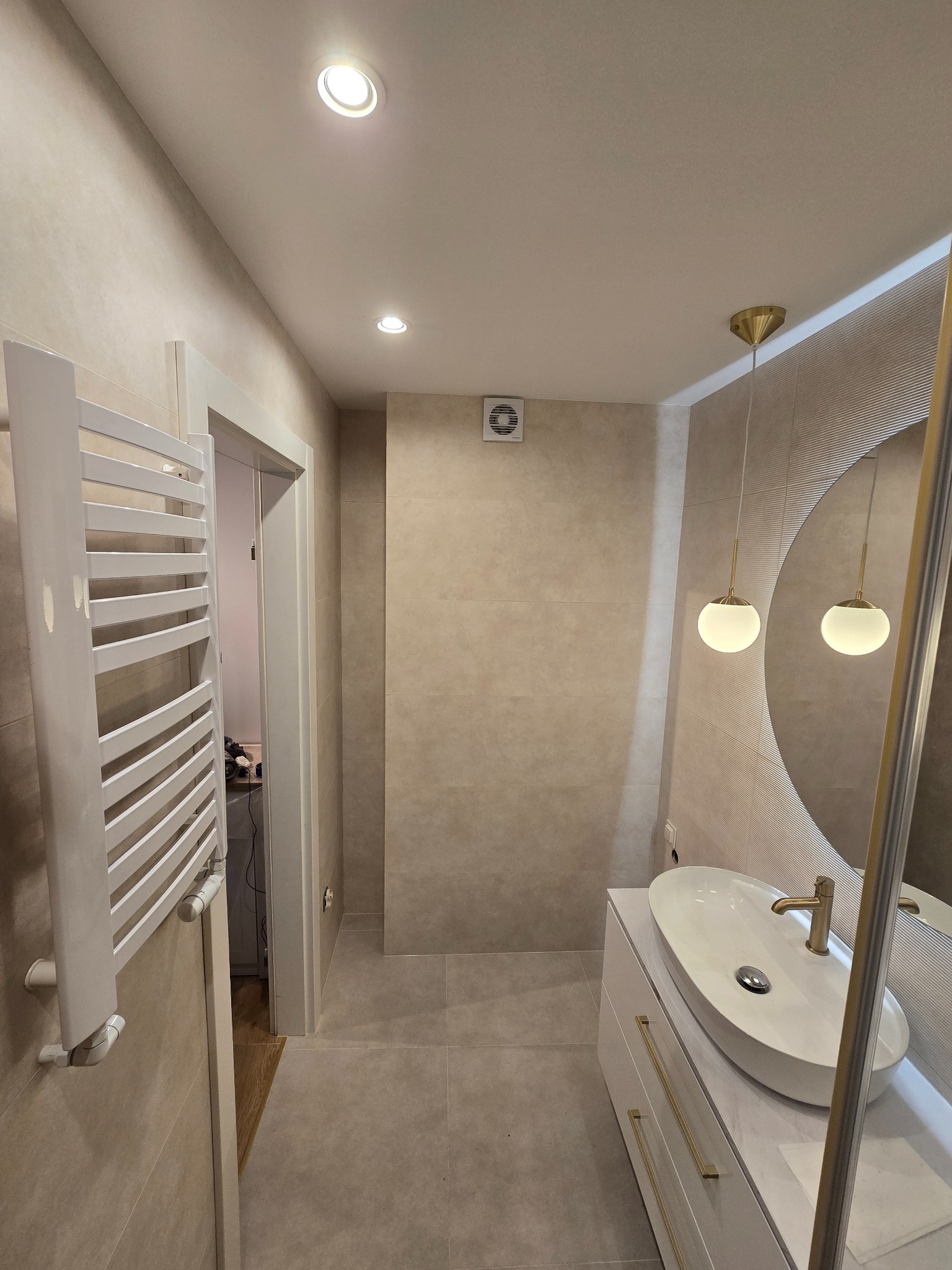 Beżowa łazienka po remoncie: owalne lustro z oświetleniem, biała umywalka na szafce z złotymi uchwytami, grzejnik drabinkowy i płytki w odcieniach beżu.